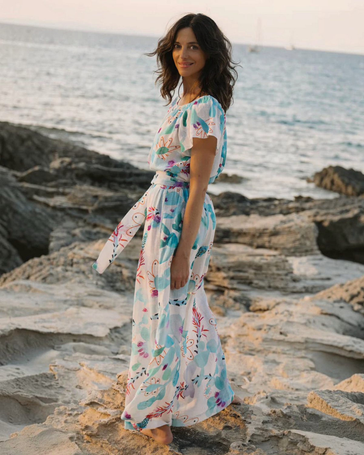 Birdie by Nina Liddle Design Dani jumpsuit in white cotton linen with blue and orange floral print on a rocky beach